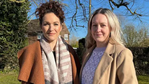 Amira Stallion and Amber Bradly stand side by side with a hedge behind them. Amira is wearing an orange coat and patterned scarf. Her brown curly hair is tied in a ponytail. Amber is wearing a cream coat and has blonde mid length hair.