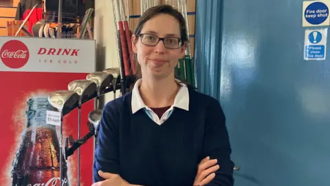 A woman is standing facing the camera with both arms folded across the front of the body. She is wearing a dark, long-sleeved sweater layered over a light-coloured collared shirt. The person is also wearing rectangular eyeglasses. On the left side of the image, there is a tall red refrigerator branded with Coca‑Cola. Behind the person and the refrigerator, wall-mounted slat panels hold various items including several golf clubs.