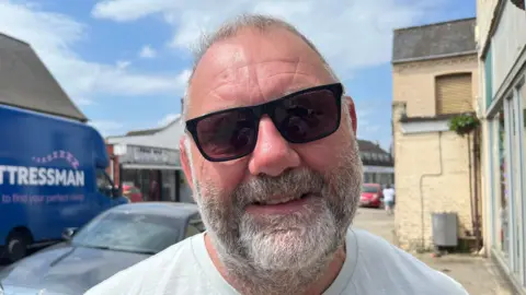 John Devine/BBC Brian Brian-Platt looking at the camera. He has short grey hair and a beard and is wearing sunglasses and a white T-shirt.