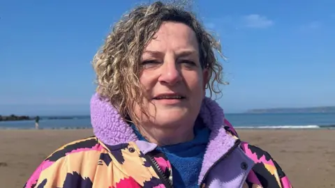 Lucy Bennett with blonde curly hair wearing a purple and pink patterned dry robe