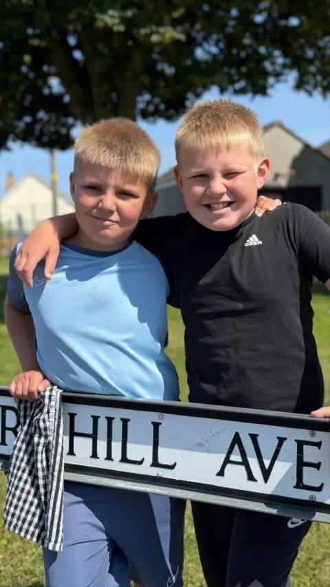 Two brothers - Jonah and Eli - stand side by side with their arms around each other smiling at the camera while they stand over a street sign. They both have short, blond hair. Jonah on the left holds a black and white checkered tea towel and is wearing a blue shirt. Eli on the right is wearing a black t-shirt and a pair of black trousers.