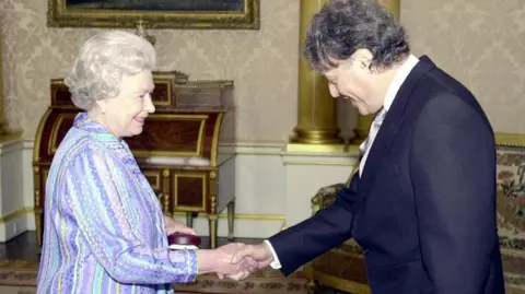 PA Media An elderly woman in a blouse with pale blue and purple stripes smiles and shakes hands with a middle-aged man who is wearing a dark suit and has grey hair.