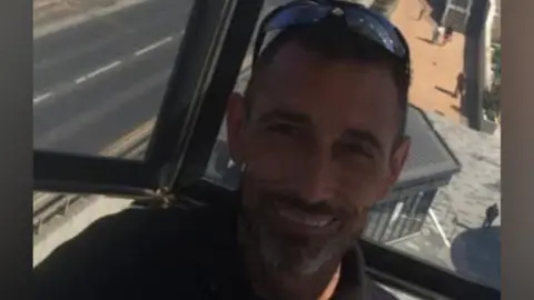 A smiling Adam Lucas has short hair and grey beard and has sunglasses balanced on his head.