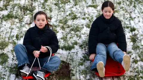 PA Media Two girls in red sleighs riding down a snow covered hill. They are both wearing jeans and black hoodies. Snow is sitting on the grass.