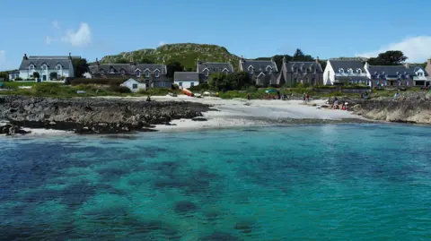 Getty Images A row of small houses on a rocky shoreline with a small beach and a turquoise-coloured sea. 