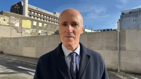A man wearing a suit and tie standing in front of a boarded up wall with a few buildings behind it in vision and blue skies behind. A road is in front of the wall.