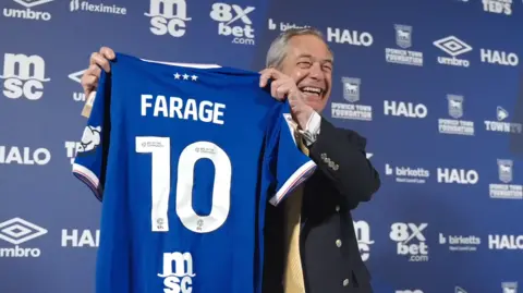Nigel Farage poses with a blue Ipswich Town home shirt with 'FARAGE 10' on the back. He is standing in front of a blue advertising wall with various sponsors' logos on it. He is looking away from the camera and laughing.
