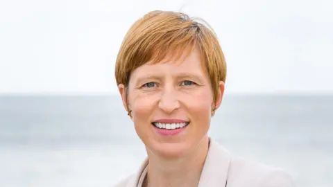 Clare Morley. She has short reddish brown hair and blue eyes. She is wearing a white blazer and is smiling at the camera.