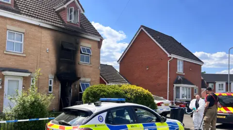 A firefighter and a forensic officer wearing a white suit stand to the right of a property in a residential street that has been damaged in a fire. The front door semi-detached property has been destroyed and large black streak covers the bricks and the window above the door. There us a police car parked in front of the house and blue police tape has cordoned off the house and the driveway on the right of the property where a white car is parked.