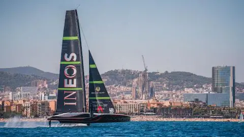 Ineos Britannia The Ineos Britannia yacht, with two triangular black and green sails and a black hull, at sea with Barcelona behind.