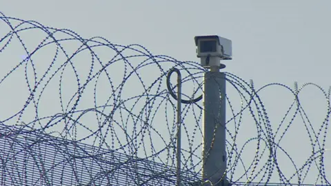The image shows the top section of a high‑security perimeter fence covered in tightly coiled razor wire. Above the fence is a mounted CCTV camera on a metal pole.