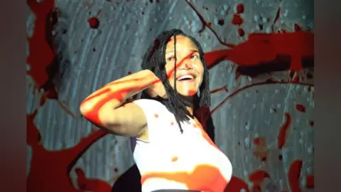 Women of the World/Vitalia Jackie Ellis in a stylised image with splashed of red across the screen, she has one hand behind her head and is look upwards.