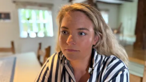 Hannah is looking off to the left of the screen. She has blonde wavy hair that is pushed back behind her ears. She is wearing a blue and white striped shirt. Behind her the background is blurred. You can see the dining room table that has a blue runner down the middle. There is a window behind her and a blurred picture of her dog Betsy on the wall.