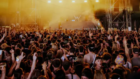 Alastair Brookes A large crowd of young people cheer and wave their arms while facing a stage inside a marquee. The photograph is taken from the back of the crowd looking towards the stage where a DJ in a cowboy hat has their arms raised.
