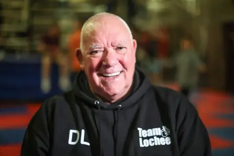 Derek Lynch looks into the camera smiling while sitting in a boxing club, which is blurred in the background. He is wearing a black hoodie that says 'Team Lochee' in white writing. He is balding with white hair.