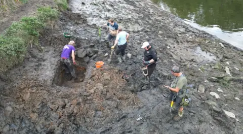 Durham University and Gary Bankhead Five volunteers digging a trench in the mud at the river bank. One person is standing in the hole and is digging with a spade. Two others also hold spades while the two remaining men are holding metal detectors.