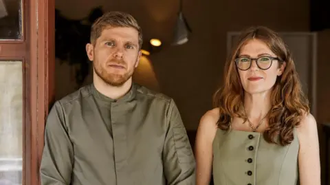 Safia Sarkarchi Kirk Haworth, a man with light hair and facial hair, wearing an olive green shirt, standing alongside Keeley Haworth, a woman with long wavy ginger hair, glasses, wearing an olive green dress.