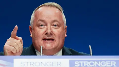 Darren Millar pointing in the air with his right hand, speaking in front of two small microphones at a lecturn that has the word "stronger" written in capital letters. He is stood in front of a blue background at what appears to be a Tory party conference.