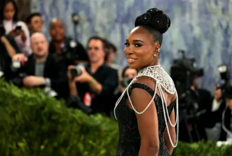 Reuters Venus Williams poses during the Met Gala, an annual fundraising gala held for the benefit of the Metropolitan Museum of Art's Costume Institute.  Williams wears a black, sparkling gown with a flared train, but the star of the outfit is her large silver neck adornment.