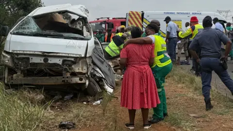 A rescue worker in a high-vis jacket comforts a woman in a red dress. A damaged white bus is situated ahead of them.