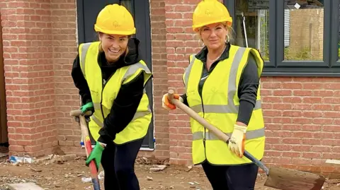 Prima Chloe Radcliffe and her mum Cordelia Hepworth, holding shovels and wearing hi-vis jackets and hardhats, stand in front of one of the partly built homes on an estate.