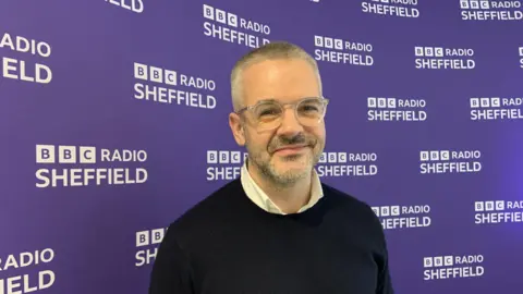 A man with close cropped grey hair is wearing glasses, a white shirt and a black jumper. He is standing in front of a purple wall with the words BBC Radio Sheffield in white