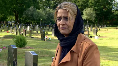 A woman standing looking into the distance looking upset. She is wearing a brown jacket and has a black scarf around her head. 