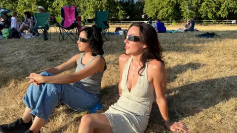 Two woman are sat down on grass with special eclipse glasses looking at the sun