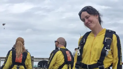 Scott Armstrong Three people outdoors are wearing bright yellow jumpsuits with black straps around them. Two, on the left, are watching a parachutist in the sky. Belinda Taylor, on the right, is looking at the camera with a small smile and her head tilted slightly to one side. She has brown hair and is wearing goggles on her head