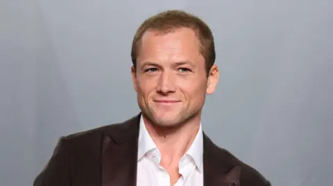 Egerton, a man in his late 30s wearing a white shirt and a brown suit jacket. There's a grey background behind him.  