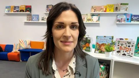 Helena Thompson is sitting in the school's library. A number of children's books are on display behind her. 