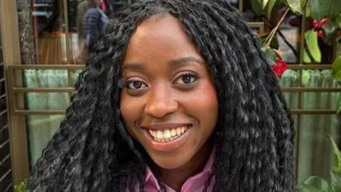 Minah Shannon has long black wavy hair and is wearing a purple shirt. She is smiling at the camera.