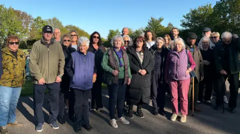 A line of 21 men and women standing on a road at a cemetery. Most look in their 60s and 70s, though some are younger. They have serious expressions on their faces. Gravestones and trees can be seen in the background under a blue sky.