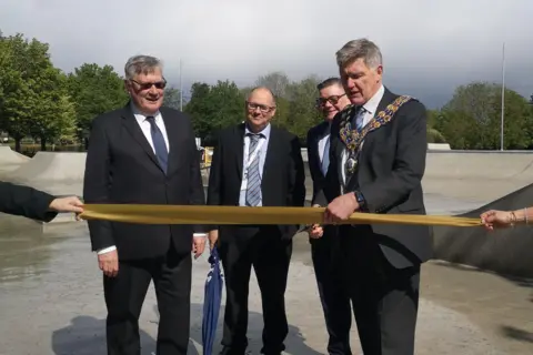 Maldon District Council Four men in black or navy blue suits, and ties, are standing on the concrete of a skate park. One of them is cutting a yellow ribbon with scissors. It is cloudy above.