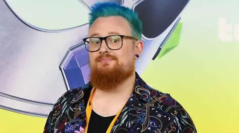 Warwick, a man with dyed blue hair and a ginger beard, looks into the camera. She wears glasses and has a pierced nose