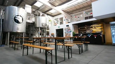 Small Beer Brewery A taproom where you can see the tanks for the brewery in the background. Bunting flags decorate the room with large wooden benches to sit upon. A bar can be see in the background.