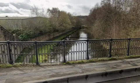 A view from a bridge with black railings across it. The view is of a canal stretching into the distance with a footpath, brick wall and trees alongside it on both sides.