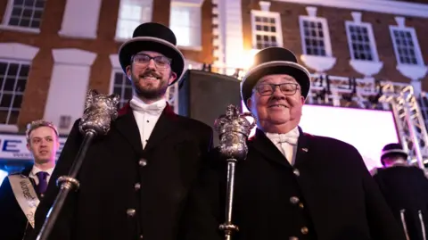 Two men wearing Victorian-era black suits and black top hats are pictured holding metal staffs with a royal crown at the top. 