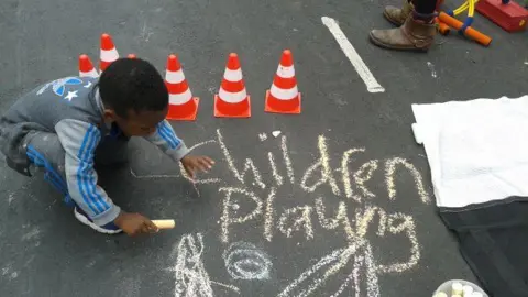 Playing Out A young boy chalks the words "children playing" on the street. Some small traffic cones used in children's games sit close by. 