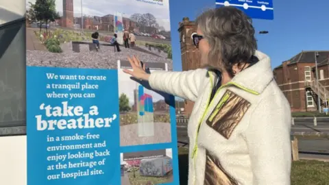 Paula is wearing a cream and gold fleece with a neon yellow zip, she is looking at a display poster showing how the planned garden will look. 