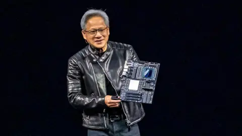 Jensen Huang wearing a black leather jacket and jeans, standing on a stage holding a circuit board