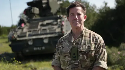 Lee Durant/BBC Major General Tom Bewick, who is middle-aged and has short brown hair, is seen standing in front of an army tank