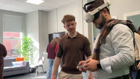 A man in a striped shirt is using a VR headset and hand controls. He is standing in a room which is brightly lit by a window. A man wearing a brown shirt is observing him. Other people can be seen in the background.