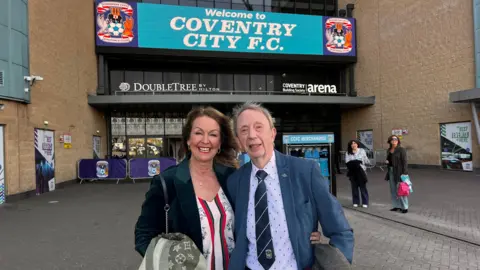 Julie White A woman with brown hair is smiling and is wearing a black jacket and white, green and red top. She has her arm around her father who has grey hair and a blue suit jacker, with a white shirt and black tie