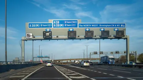 A motorway with six lanes - two are going to the left and the others are heading straight. There is a large gantry with road signs that are blue. One reads "the north west, birmingham north and west, m6"