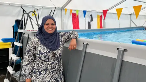 A woman in a patterned dress and headscarf stands beside an above-ground swimming pool inside a large tent, decorated with colourful triangular bunting. A metal ladder is positioned at the pool’s edge
