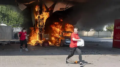 Two women in red tops run as flames rise from a car and fuel dispenser following a drone attack by Russia on a petrol station near the city centre of Kharkiv on 3 July