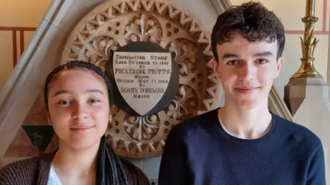 Chanel (left), who is wearing a brown cardigan and white T-shirt stands beside Thomas is wearing a blue jumper
