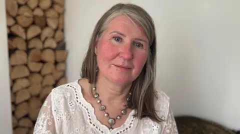 Deirdre McCanney has a neutral expression on her face as she sits in her front room. Behind her is a wood feature but it is blurred. Deirdre has shoulder-length light brown and greying hair. She is wearing a white lace top with some eyelets.