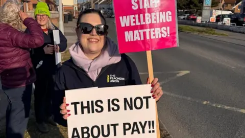 NASUWT South West NASUWT South West members stood outside the blue gates of Budmouth Academy with their banners. One female teacher is stood at the front of the image with two clearly visible signs reading "Staff wellbeing matters!" and "This is not about pay".